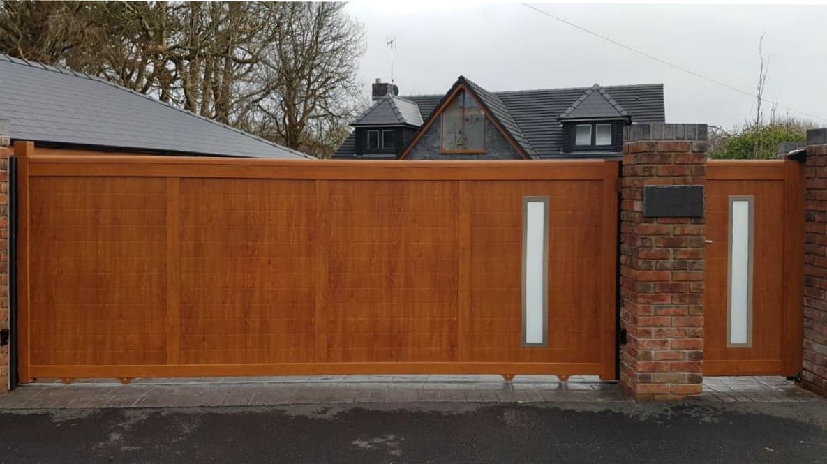 Wood-effect aluminium driveway gate with vertical panels, complemented by rectangular white inserts, set against a backdrop of a modern home with slate roofing.